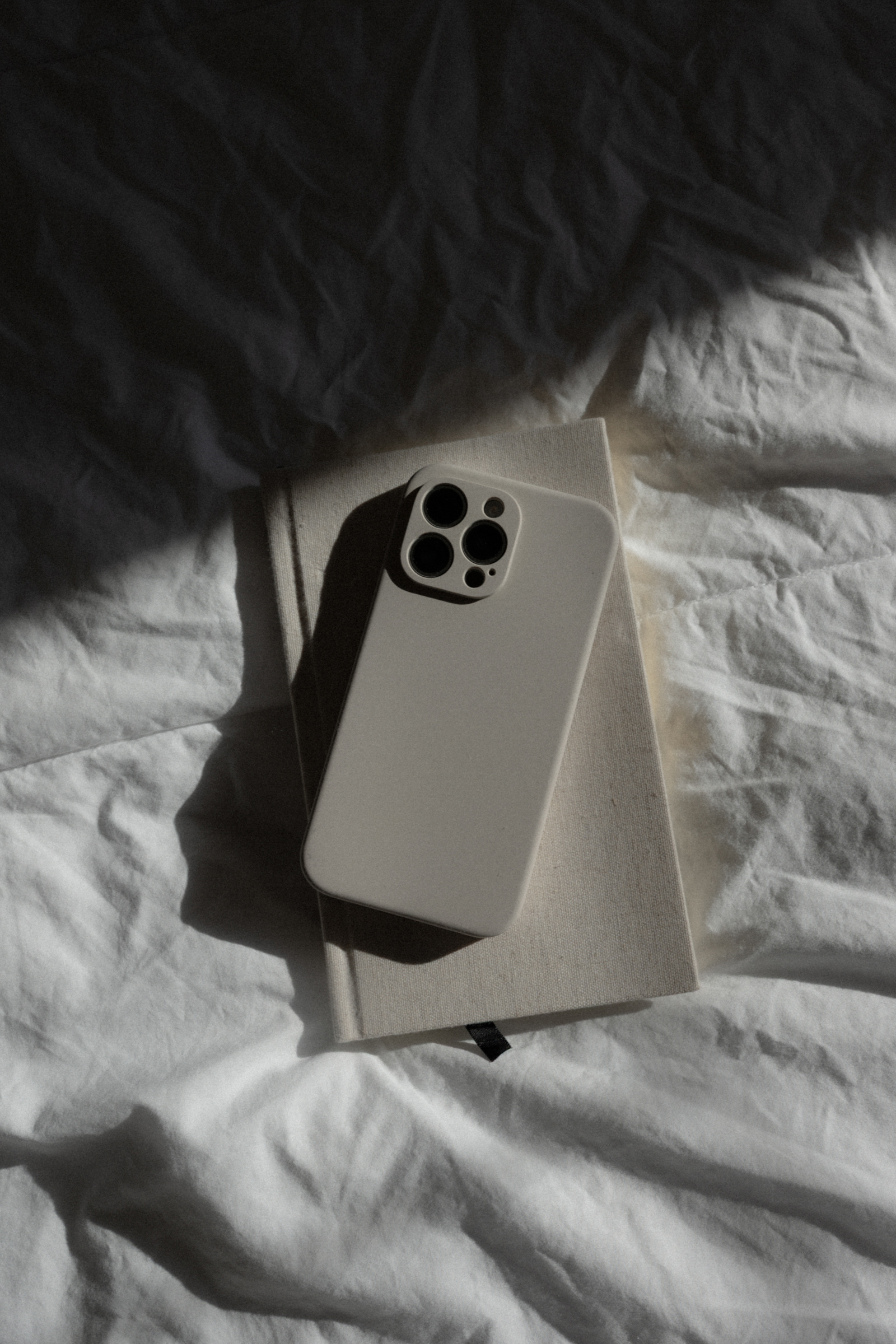 Neutral smartphone resting on a fabric-covered notebook on white bedding with dramatic shadows – True North Visionaires